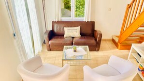 A living room with a brown leather sofa, two white armchairs, a glass coffee table with a small plant, and a wooden staircase in the background.