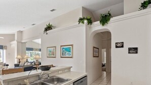 A modern kitchen with a double sink, dishwasher, and a countertop with a bowl. There are framed pictures on the wall and a plant on the ceiling. - In Orlando area, large Family Friendly Home - Close to Disney (Clermont)
