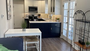 A compact kitchen with white cabinets, a microwave, and a sink. There's a dining table with a chair, a green cushion, and a white crocheted bag. A metal rack with glassware is visible. - Completely updated home! -Close to hospitals, UD, Wright Patt Air Force Base. (Kettering)