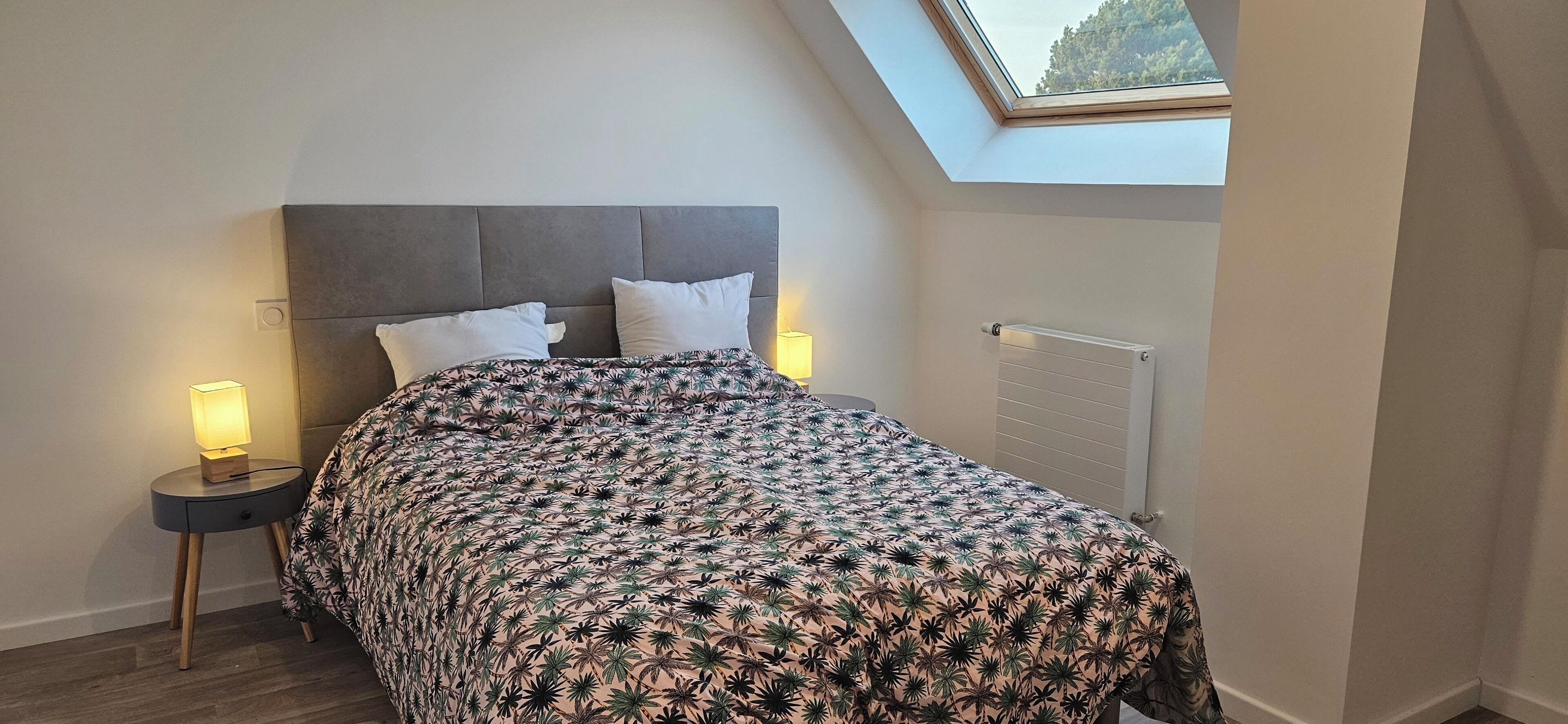 Quarto com cama, duas luminárias de cabeceira, radiador e claraboia.