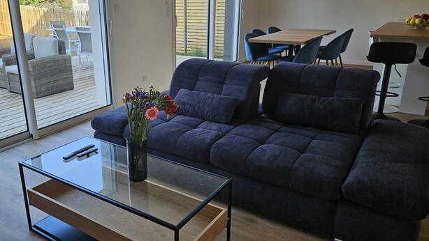 Ein modernes Wohnzimmer mit einem dunkelblauen Sofa, einem Glastisch und einer Vase mit Blumen. Ein Schiebetürfenster führt auf eine Terrasse mit Gartenmöbeln.