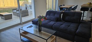 A modern living room with a dark blue sofa, a glass coffee table, and a vase with flowers. There is a sliding glass door leading to a patio area with outdoor furniture. - Les Maisons de la Plage (Saint-Philibert)
