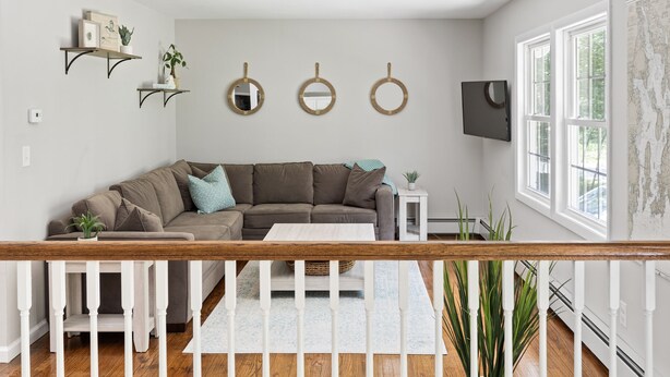 Ein Wohnzimmer mit einer Eckcouch, einem Couchtisch und einem weißen Geländer. An der Wand befinden sich Regale mit Dekoration und Pflanzen.