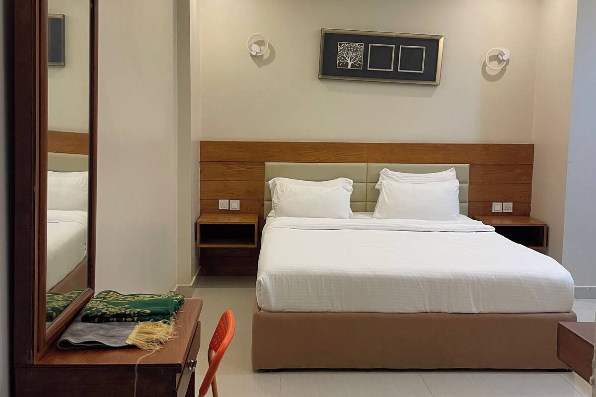 A hotel room with a large bed, a wooden headboard, a nightstand, a desk with a chair, a mirror, and a wall-mounted air conditioning unit.
