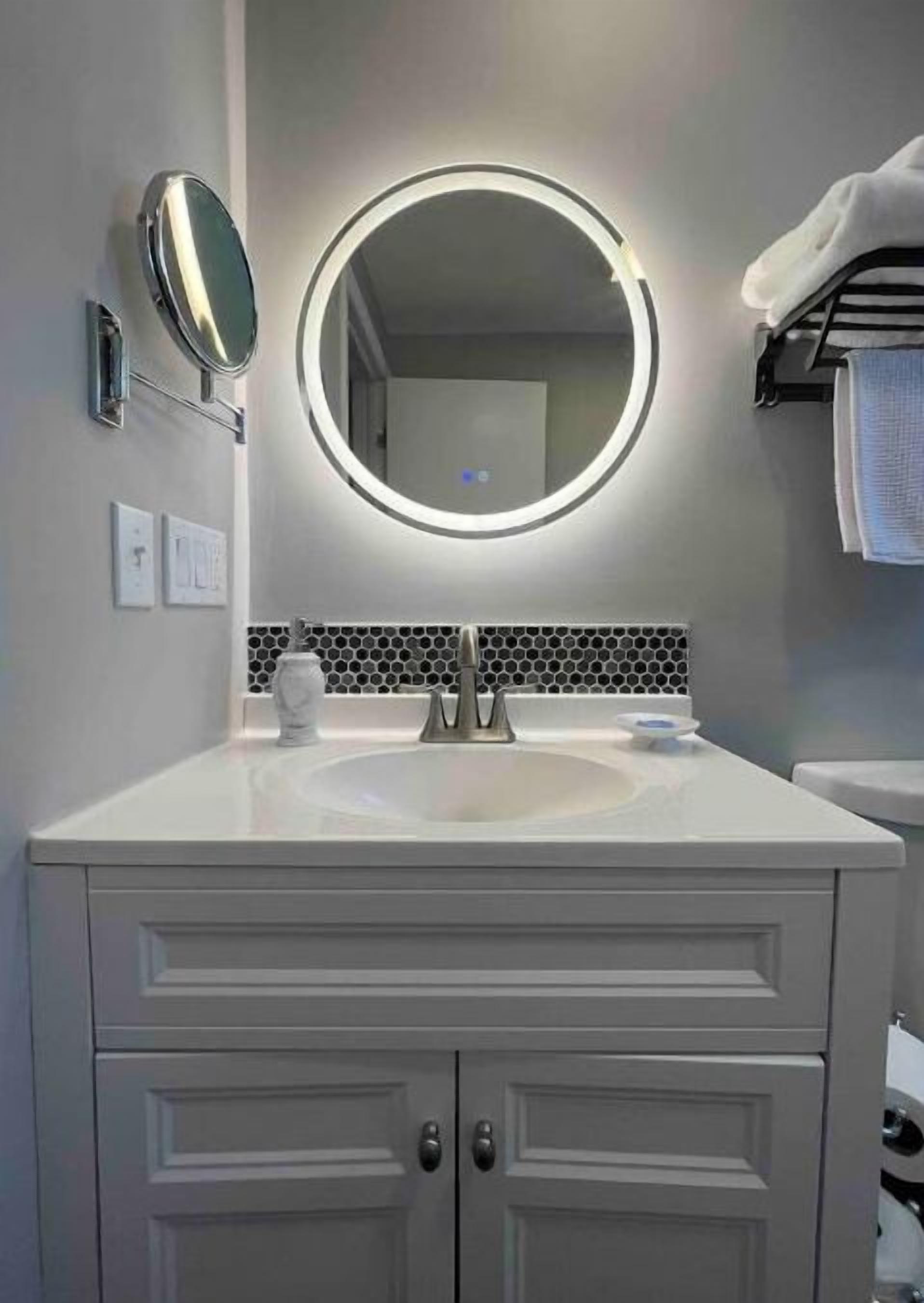 A bathroom with a white vanity, a round mirror with a light, a sink, and a tiled backsplash.