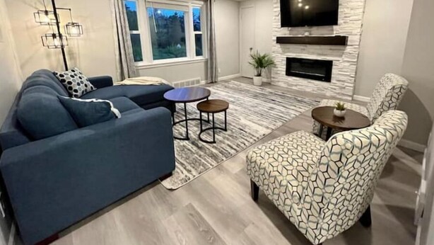 A modern living room with a sectional sofa, a patterned armchair, a small round table, and a flat-screen TV mounted on a stone wall. - Country Retreat One (North Tonawanda)