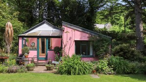 Una casa rosa con una porta e finestre verdi, immersa in un giardino curato e ricco di piante e fiori.