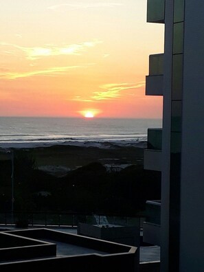 Une vue sur le coucher de soleil depuis un balcon donnant sur l’océan, avec un bâtiment moderne situé sur la droite.