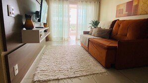 Ein Wohnzimmer mit einer Couch, einem Fernseher, einer Zimmerpflanze und Blick ins Freie durch ein Fenster.