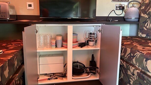 A compact kitchenette with a TV, a kettle, and a cabinet containing glasses, bowls, and cooking utensils. - Glamping Samaritan tank (Boston)