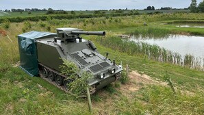A military vehicle with tracks, a mounted gun, and a protective cover, situated in a rural field with a pond and greenery in the background. - Glamping Samaritan tank (Boston)