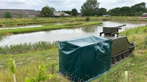 A military tank partially covered by a green tarp, situated near a body of water with grassy banks and a building in the background. - Glamping Samaritan tank (Boston)
