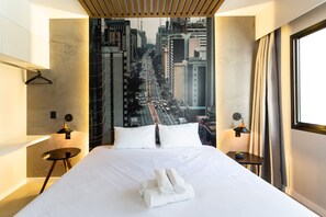 A modern hotel room with a large bed, a cityscape view, a hanging light fixture, and a small table with a lamp.