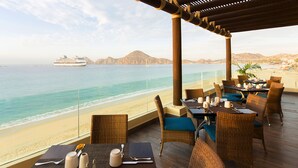 A seaside restaurant with wicker furniture, a view of the ocean, and a cruise ship in the distance. - BISBEE WEEK (Cabo San Lucas)