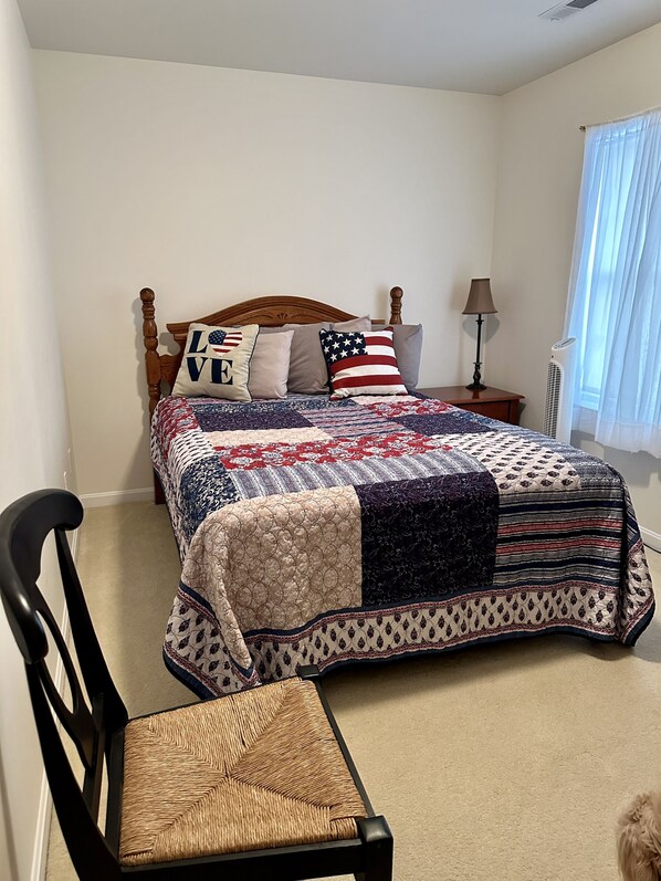 Una cama bien hecha con colcha, un cabecero de madera, una mesita de noche con lámpara y una silla con asiento de mimbre.