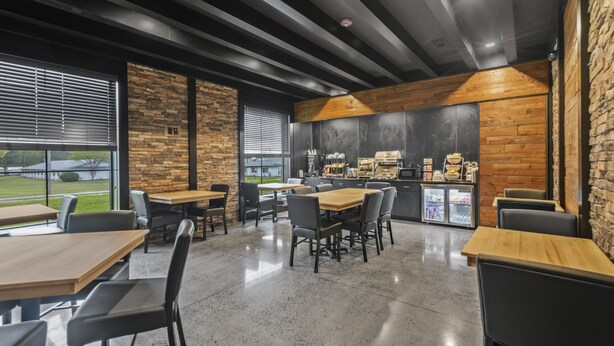 A modern restaurant with wooden tables and chairs, a kitchen area, and large windows offering a view of the outdoors. - Cobblestone Hotel & Suites - Cullman (Cullman)