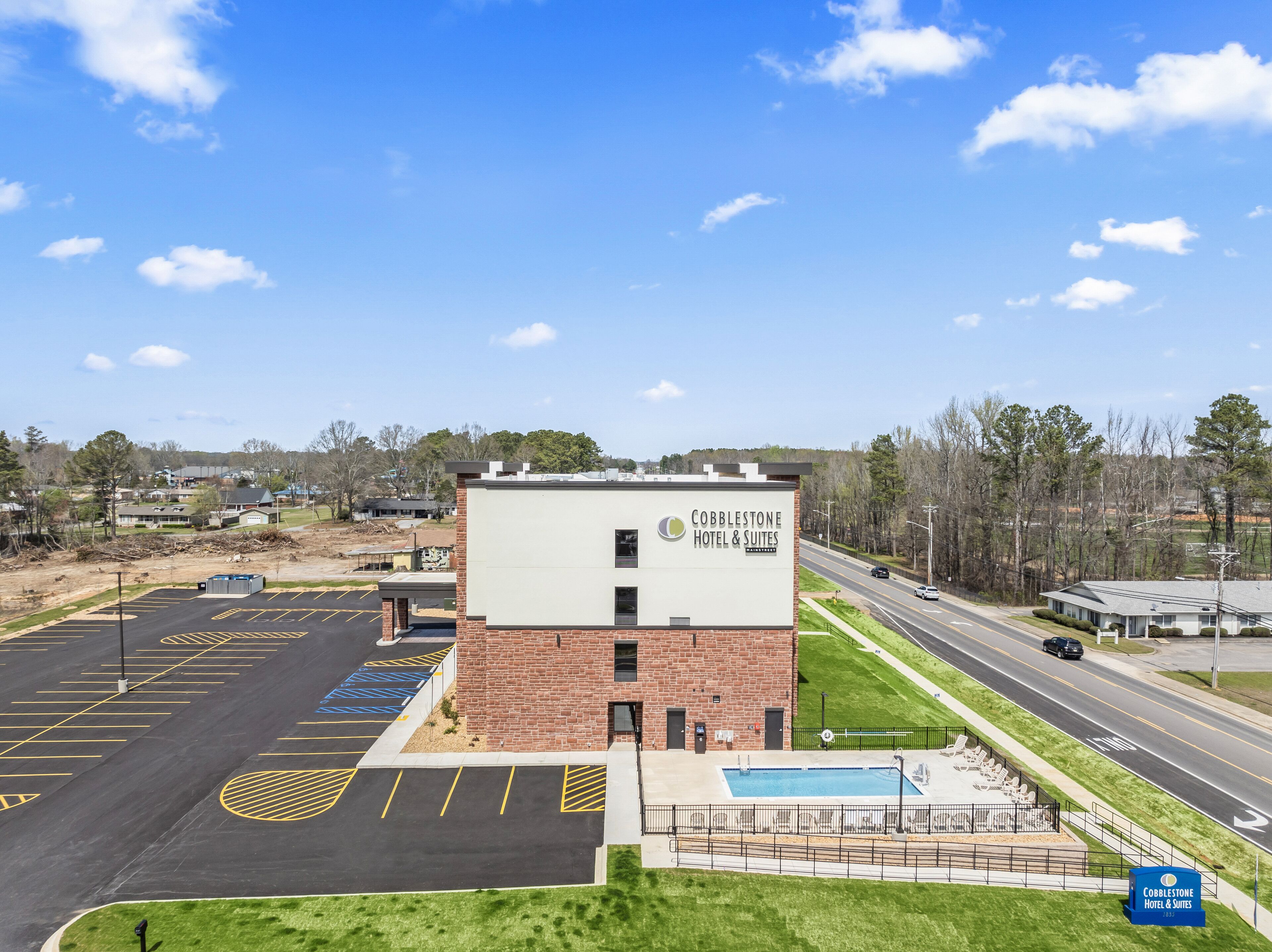 A Cobblestone Hotel & Suites with a parking lot, a swimming pool, and a road in the background.