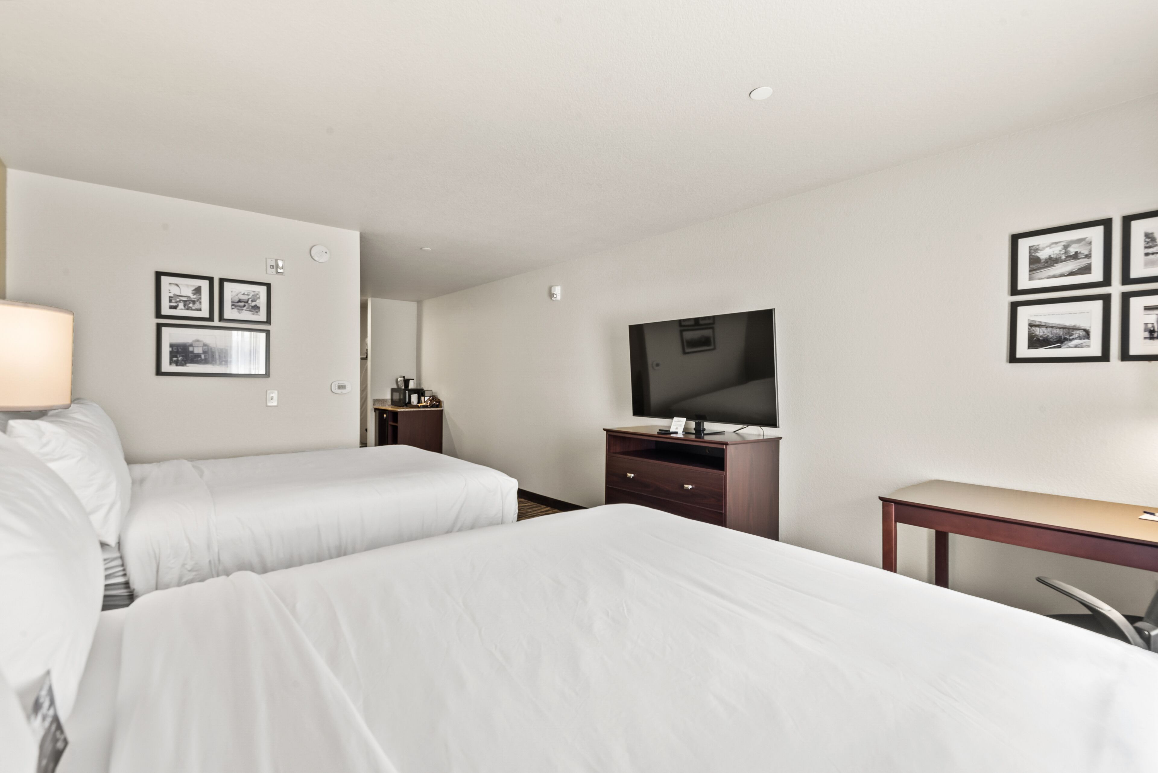 Ein Hotelzimmer mit zwei Einzelbetten, einem Schreibtisch, einem Stuhl, einem Fernseher und gerahmten Bildern an den Wänden.