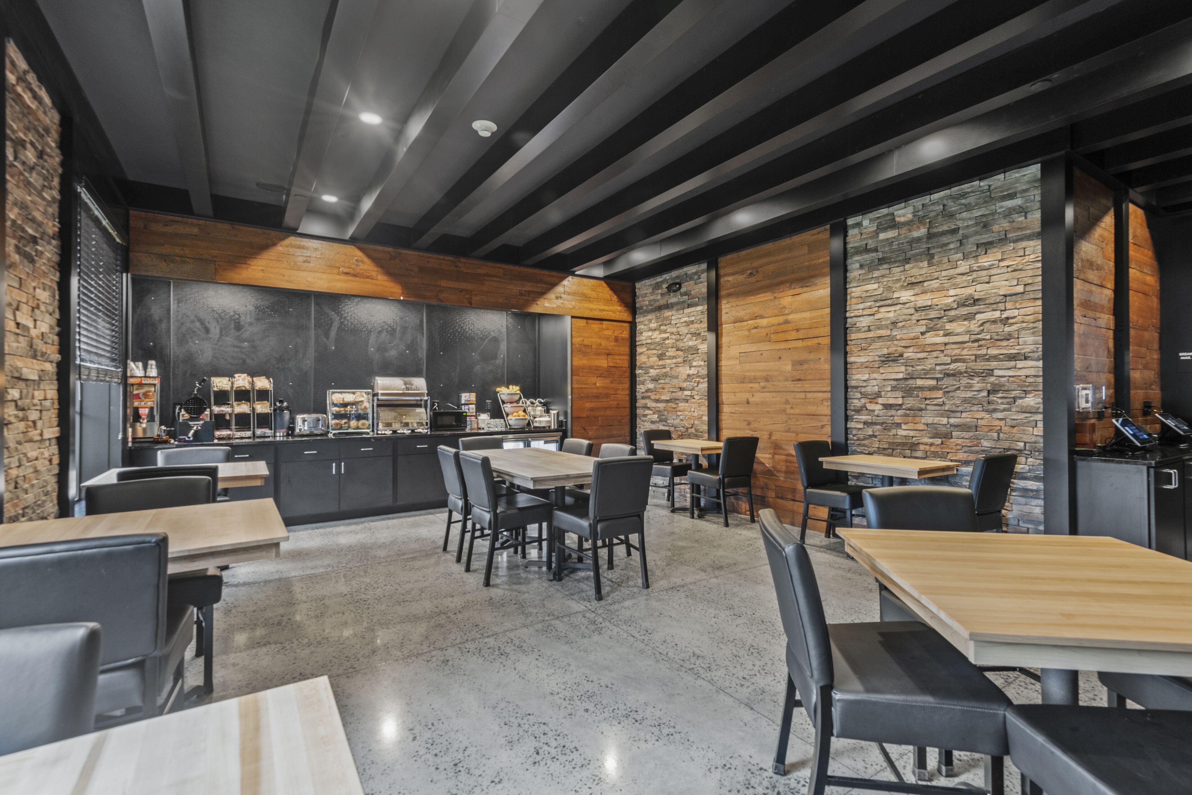 A modern restaurant with a stone wall, wooden ceiling beams, and a kitchen area visible through a glass partition.