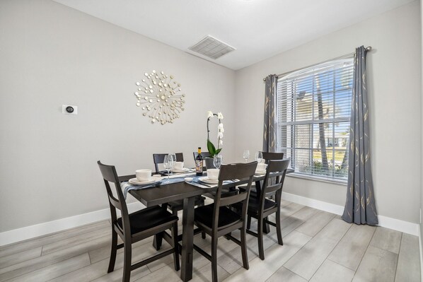 Un comedor con una mesa preparada para cuatro personas, sillas, un adorno en la pared y una ventana con cortinas.