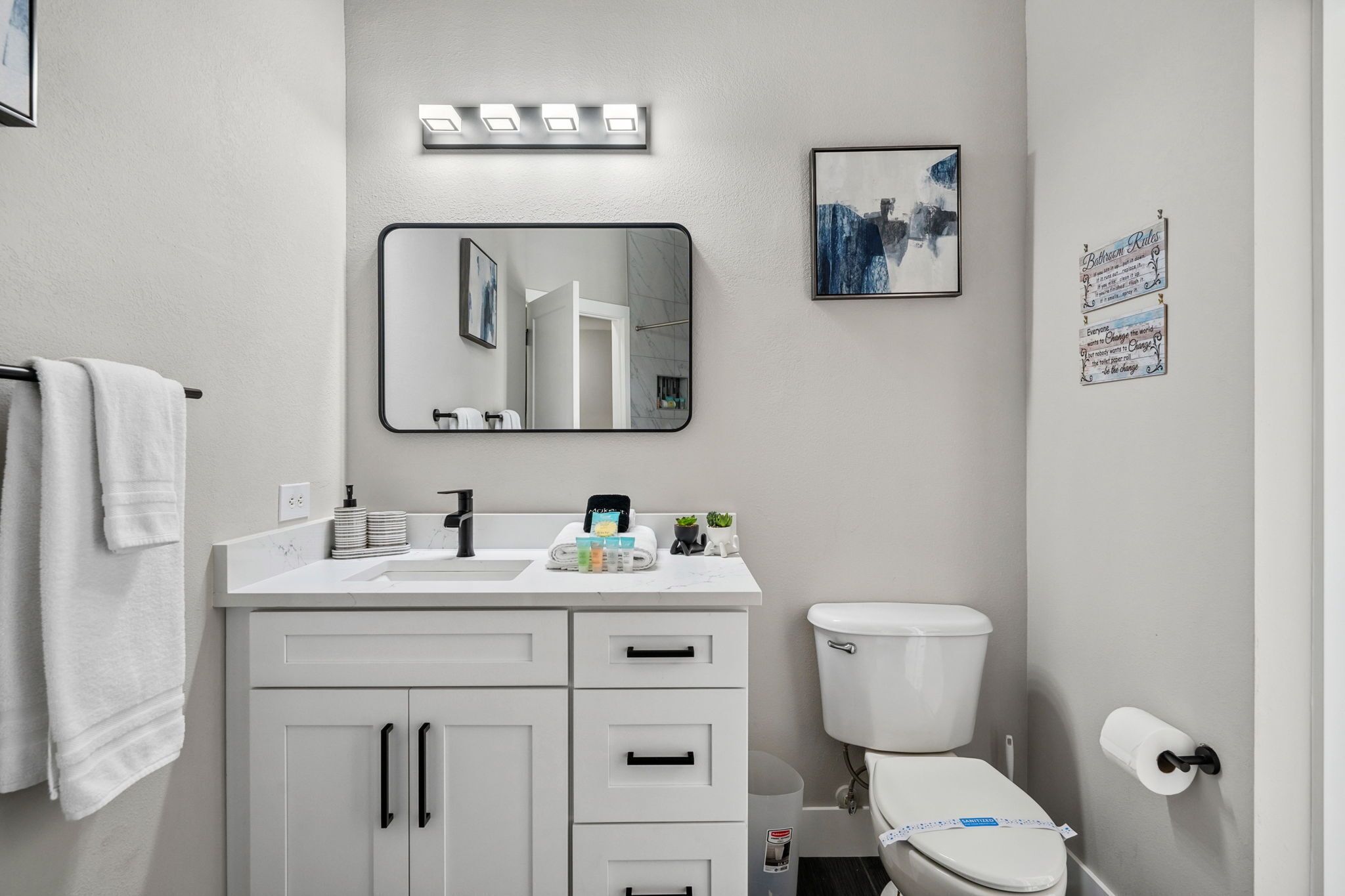 Ein modernes Badezimmer mit einem weißen Waschtisch, einem großen Spiegel, einer Toilette und einem gerahmten Bild an der Wand.