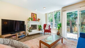 Ein Wohnzimmer mit Fernseher, Kamin, rotem Sessel und großem Fenster mit Blick auf Bäume.