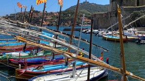 Ein Hafen mit mehreren bunten Booten, die anlegen, ein historisches Steinhaus im Hintergrund und ein klarer, blauer Himmel.