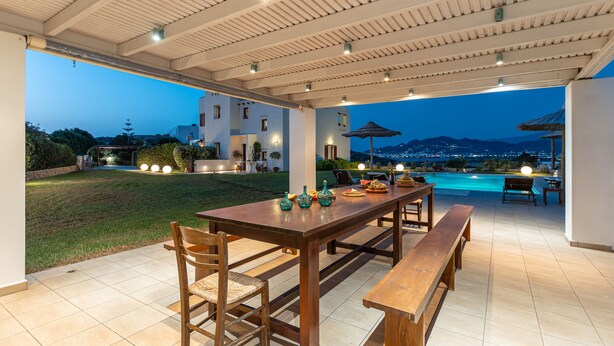 An outdoor patio with a wooden table and benches, a swimming pool, and a view of the mountains at dusk. - Villa with Private Pool & Jacuzzi Naxos (Naxos)