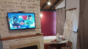 A hotel room with a brick wall, a flat-screen TV mounted above a fireplace, a dining area with a table and chairs, and neatly folded towels on the bed. - Pousada Vale do Horizonte (Camanducaia)