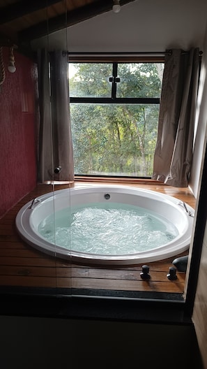 A hot tub in a room with a large window, wooden flooring, and a view of greenery outside.