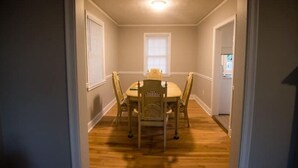 Et spiseområde med et rundt bord, fire stole, et vindue med persienner og en loftslampe.