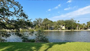 Une zone herbeuse avec une étendue d’eau, des arbres et un bâtiment en arrière-plan.