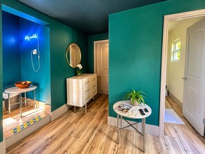 A room with teal walls, a wooden floor, a white dresser, a round mirror, a small table with a magazine and remote, and a neon sign.