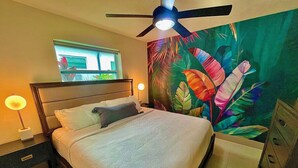 A bedroom with a bed, a ceiling fan, a nightstand with a lamp, and a wall mural.
