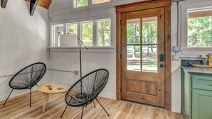 Een kamer met een houten deur, twee zwarte metalen stoelen, een klein rond tafeltje en uitzicht op bomen door de ramen.