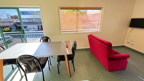 A room with a dining table, chairs, a red sofa, a window with blinds, and a TV mounted on the wall.