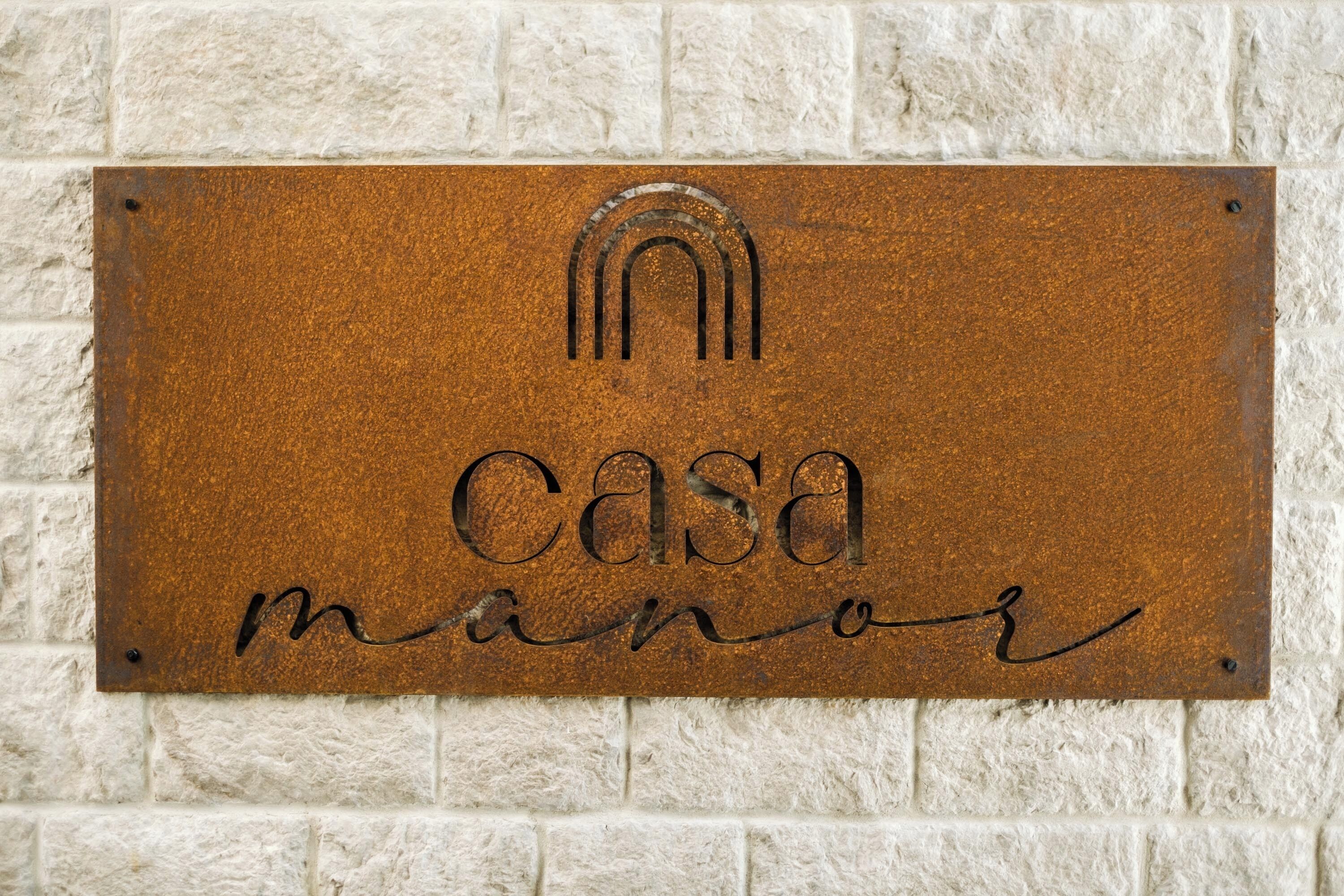 Une plaque métallique portant l’inscription « Casa manor » est fixée à un mur à texture.