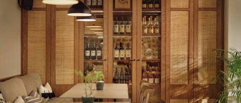 Uma área de refeições com parede de madeira, prateleiras com garrafas, mesa, cadeiras e luminárias pendentes.
