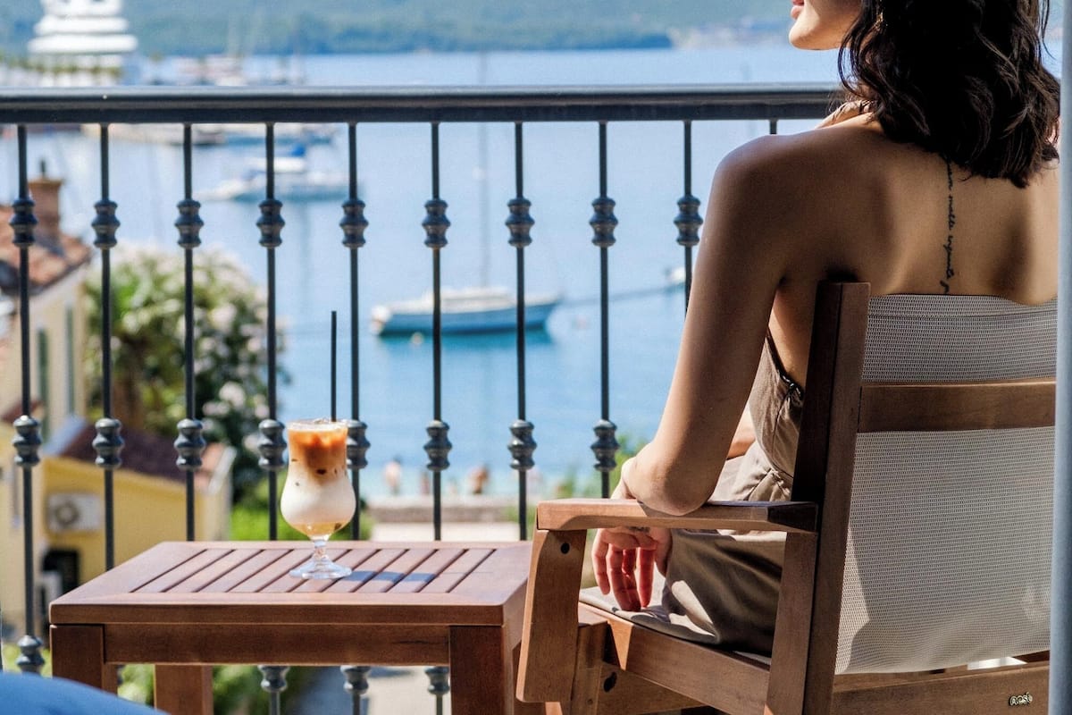 A person seated at a wooden table with a view of a harbor and mountains in the background.