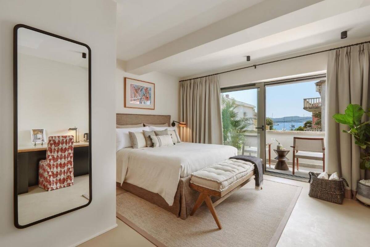A bedroom with a large bed, a bedside table, a chair, a mirror, and a view of the outside.