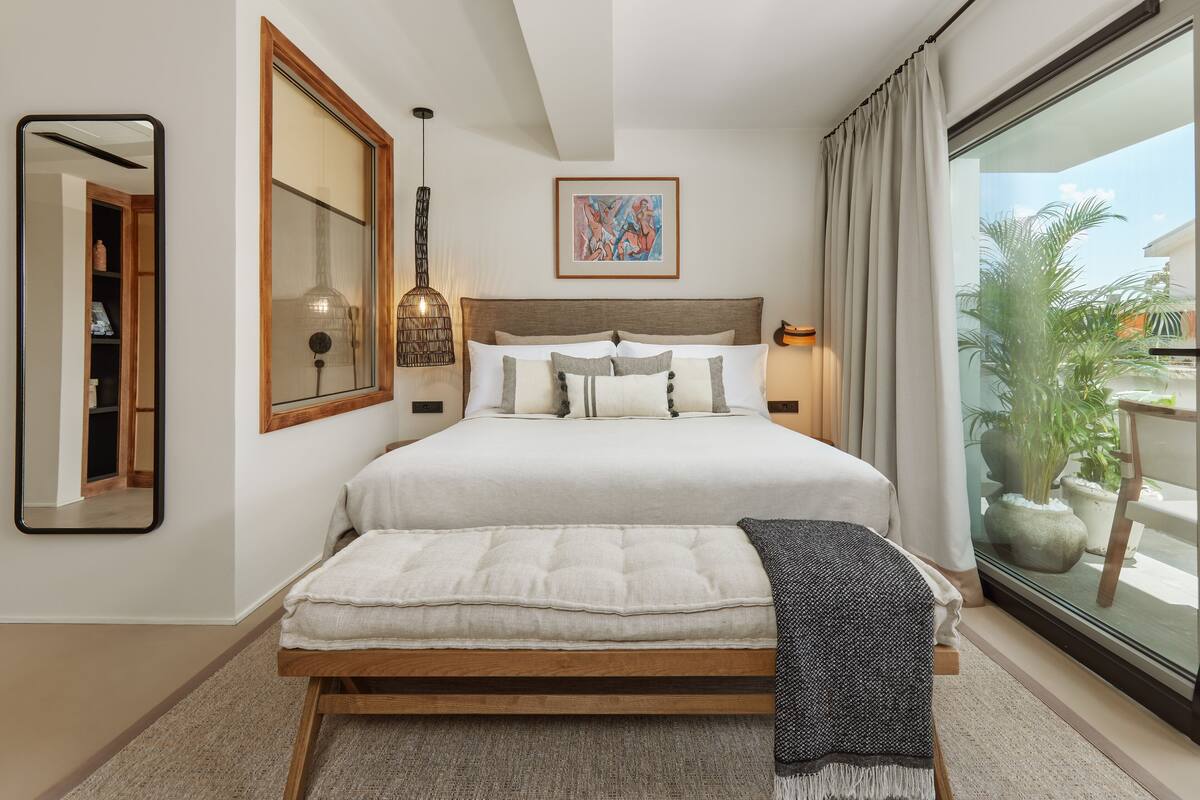 A bedroom with a large bed, a bedside table with a lamp, a window with curtains, and a view of the outdoors.