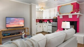 A living room with a television, a sofa, and a kitchen with white cabinets and a pink accent wall.
