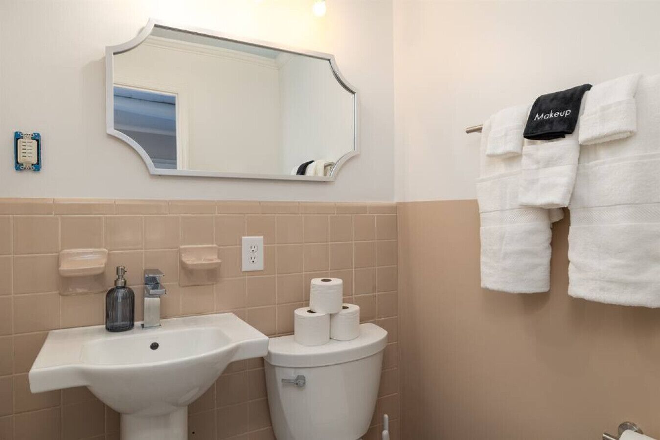 Un baño con lavamanos, inodoro y espejo. Hay toallas colgadas en la pared y un dispensador de jabón en el lavamanos.