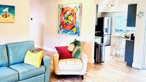 Een woonkamer met een blauwe bank, een witte fauteuil en een plafondventilator. Er hangt een schilderij aan de muur en er staat een plant op de vloer.