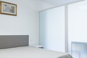 A bedroom with a bed, a bedside table, a framed picture on the wall, and a sliding glass door leading to a balcony.
