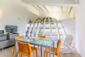 A modern dining area with a glass table, orange chairs, a white rocking chair, and a kitchen area with a microwave and TV. - GuestHost - Palermo Modern Apartment with Swimming Pool (Palermo)