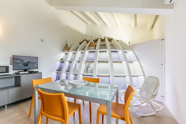 A modern dining area with a glass table, orange chairs, a white rocking chair, and a kitchen area with a microwave and TV.