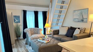 Un salon moderne avec un fauteuil à motifs gris, un canapé gris et un petit pouf sur lequel sont posés des magazines. Une échelle est adossée au mur et une lampe se trouve sur une table d’appoint.