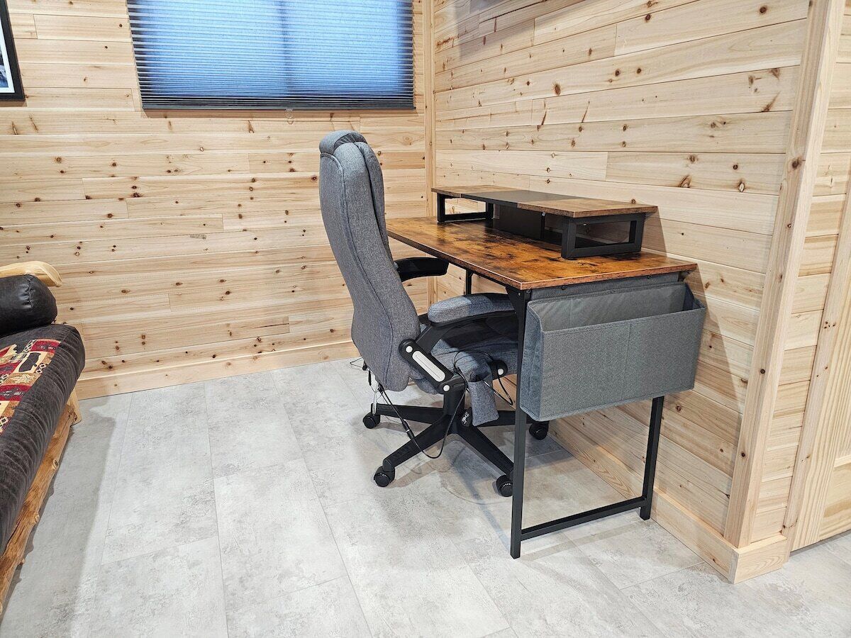 Een knus, klein kamertje met een houten wand, een grijze bureaustoel, een houten bureau en een klein raam met jaloezieën.