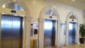 A hotel lobby with four elevators, each with a metallic finish, flanked by white columns and arches.